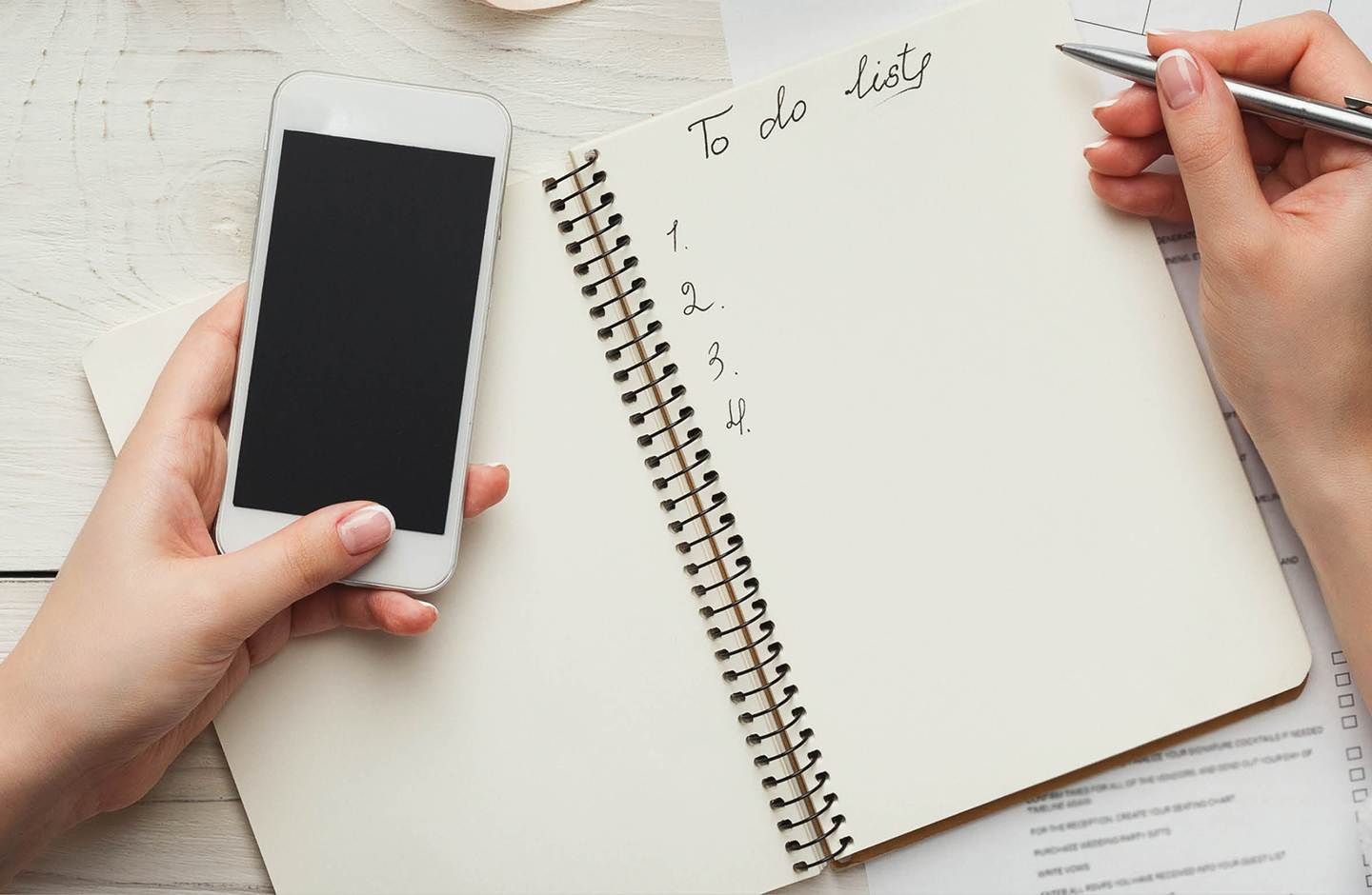 Die Hochzeits To Do Liste liegt auf dem Tisch. Es sind zwei Hände zu sehen, die einen Stift und ein Smartphone in der Hand halten