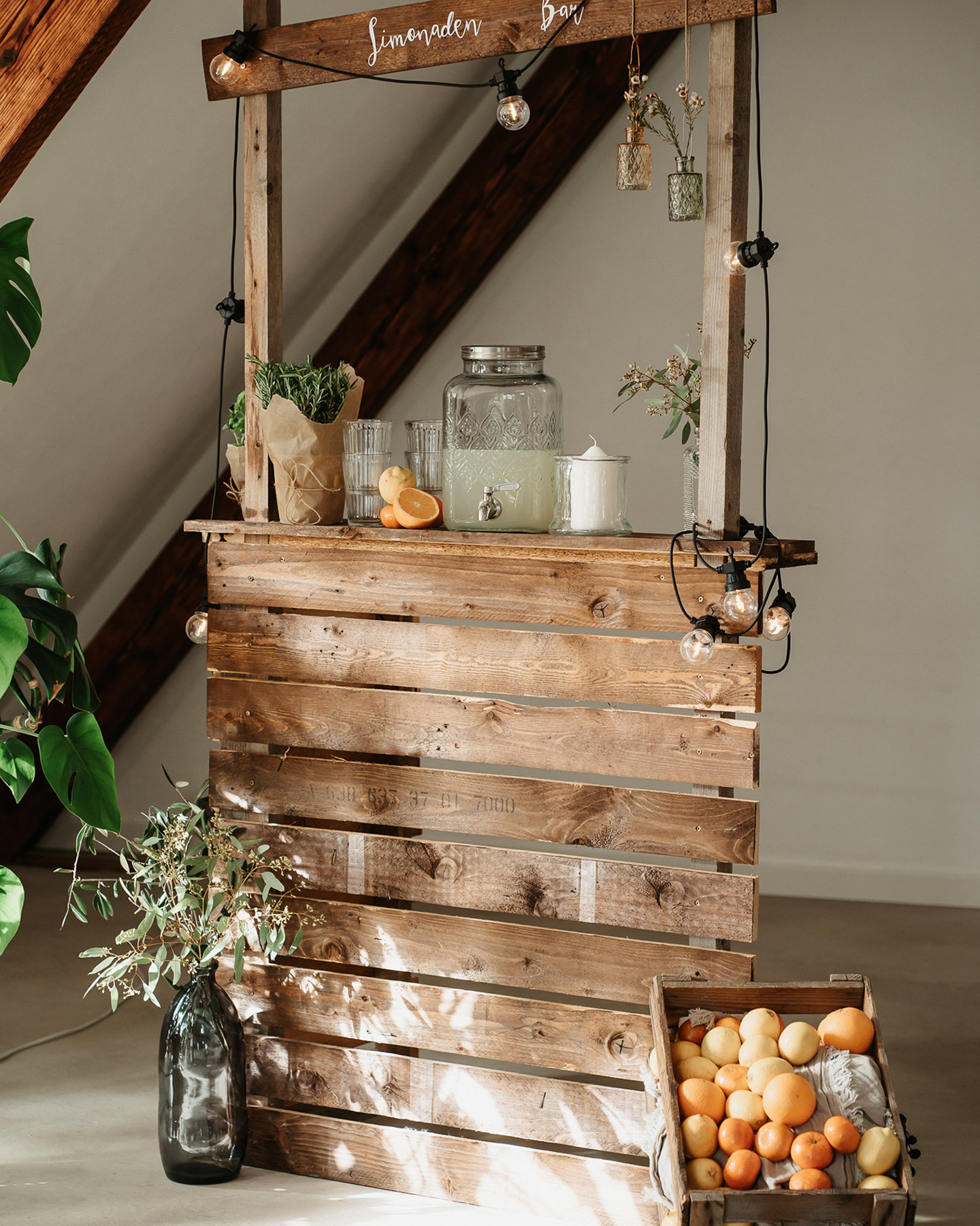 Eine rustikale Limonaden-Bar aus Holz geschmückt mit einem Korb voller Zitronen und Orangen und Olivenzweigen. Die Bar hält selbstgemachte Limonaden für die Gäste bereit.