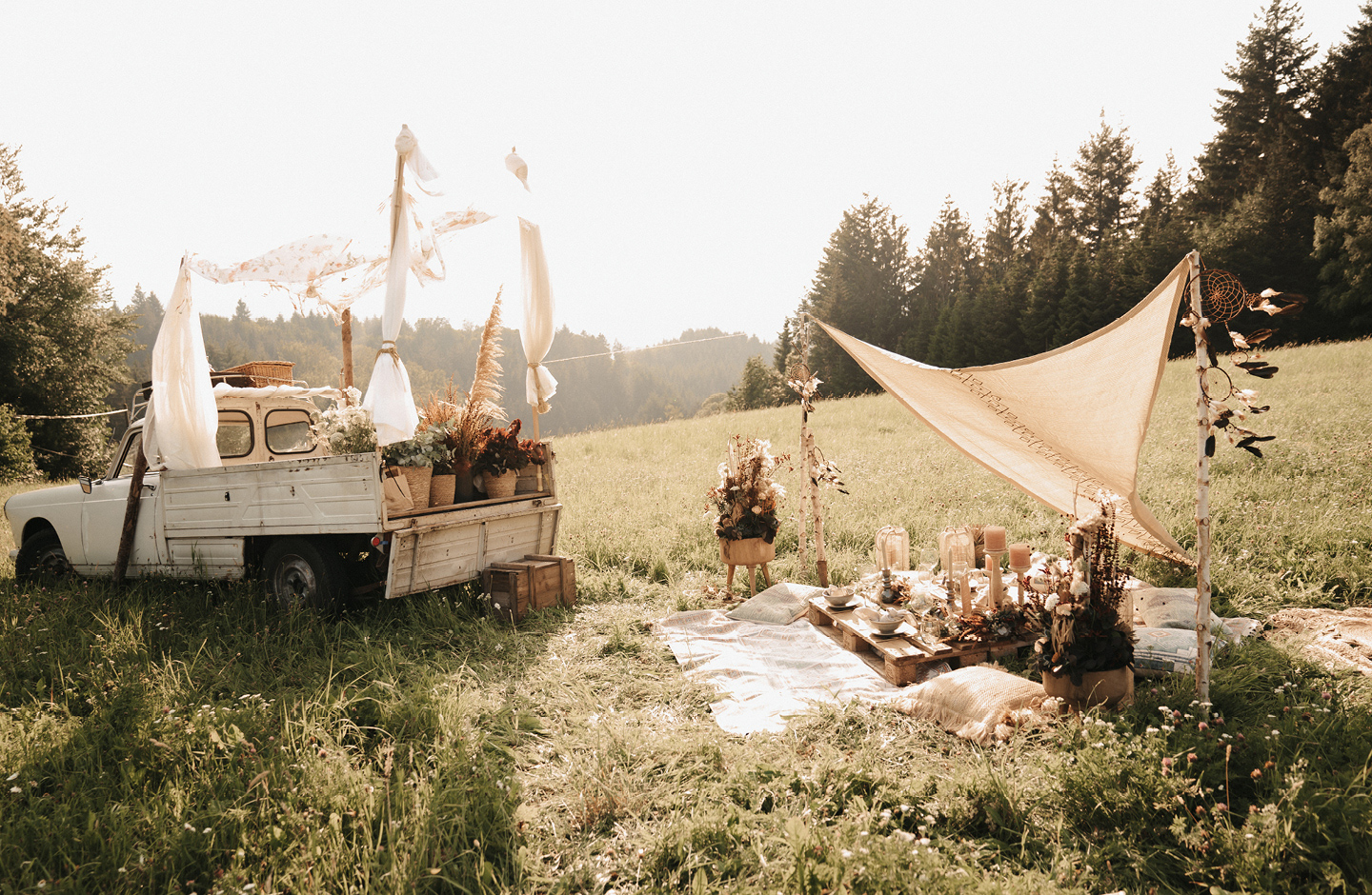 Boho Picknick ist auf grüner Wiese mit Tannen im HIntergrund aufgebaut. Eine Decke und Boho-Dekoration laden zum Verweilen ein.