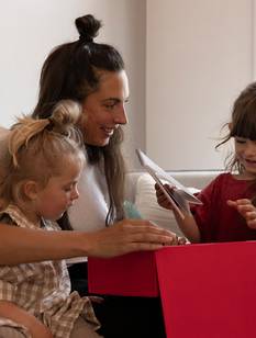 Frau auf der Couch mit 2 kleinen Mädchen öffnet ihr Weihnachtsgeschenk