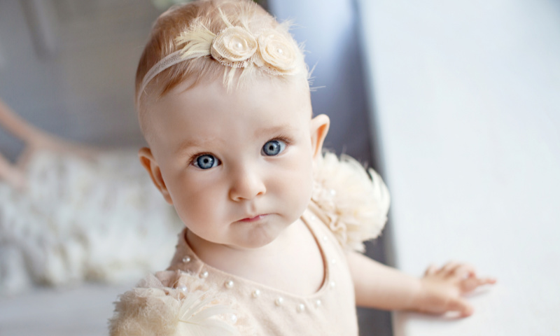 kleines Mädchen mit beige farbenen Taufkleid und Haarreif im Haar guckt in die Kamera 