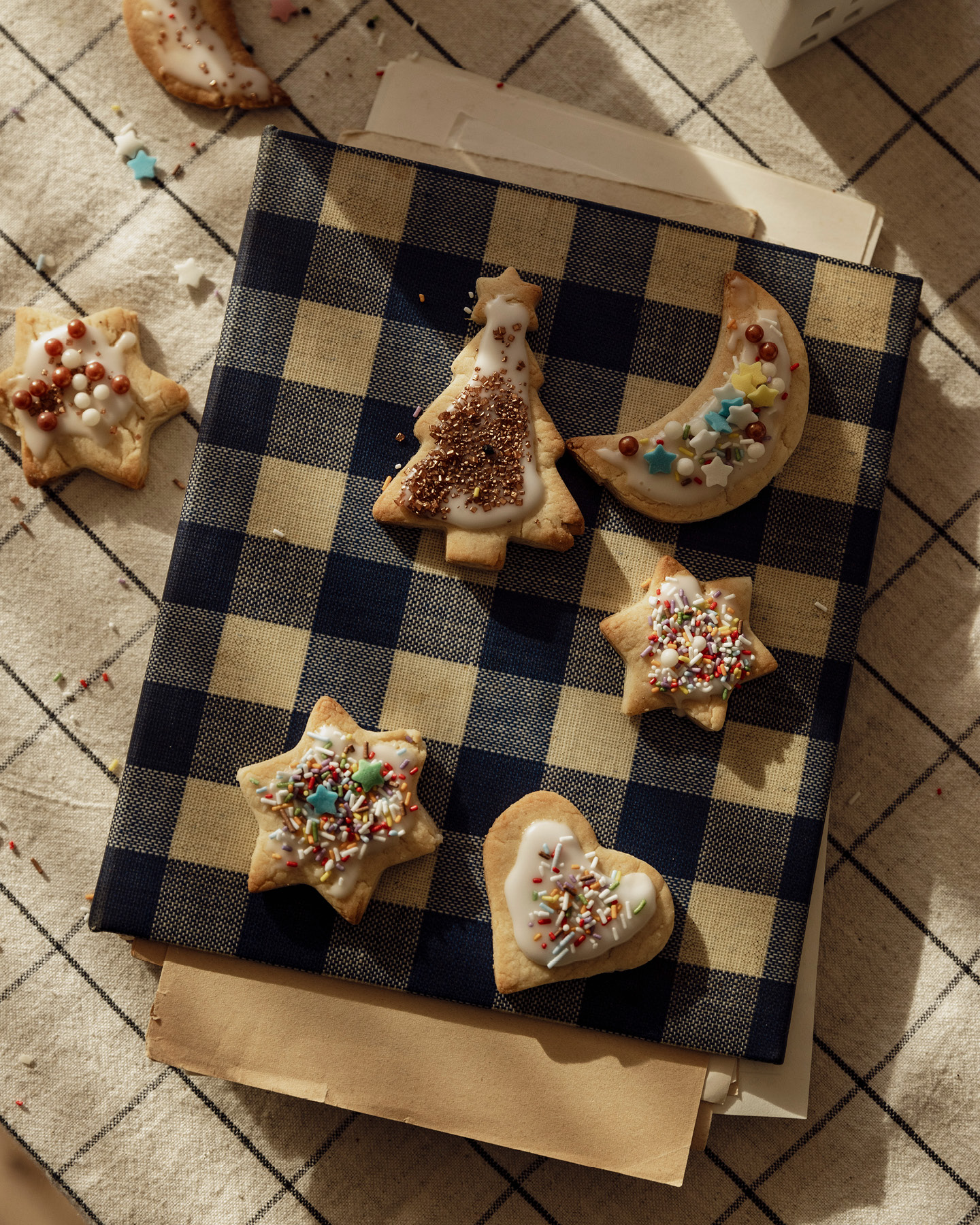 Zugeklapptes Rezeptbuch mit kariertem Einband, es schauen lose Blätter heraus, auf dem Buch sind bunte Weihnachtsplätzchen dekoriert